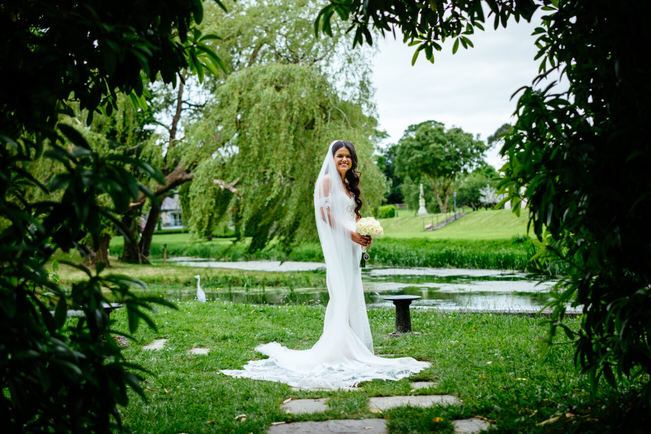 Tara and Darren’s Unforgettable Wedding at Bellingham Castle 103 A bride in a white wedding dress stands on grass near a pond at her wedding at Bellingham Castle, holding a bouquet of flowers, surrounded by green trees and lush greenery.