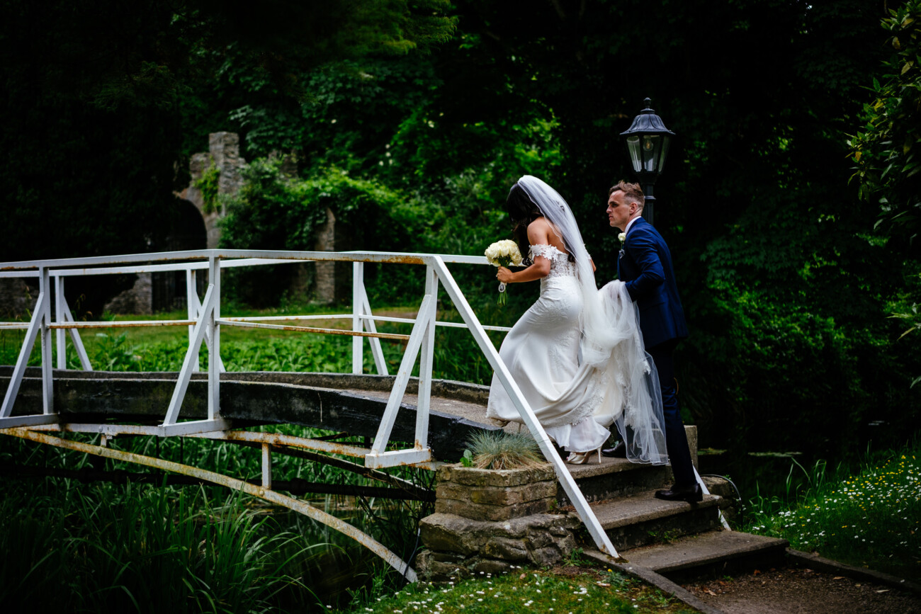 Tara and Darren’s Unforgettable Wedding at Bellingham Castle 104 A bride in a white dress and veil walks up steps on a small bridge, holding a bouquet, followed by a man in a blue suit. Lush greenery surrounds them, capturing the romance of their wedding at Bellingham Castle.