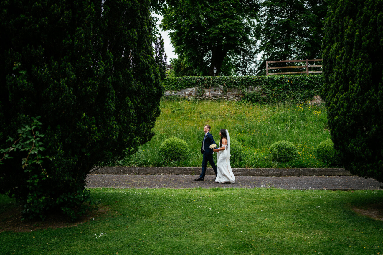 Tara and Darren’s Unforgettable Wedding at Bellingham Castle 105 A bride and groom walk together on a garden path during their wedding at Bellingham Castle, surrounded by greenery and large trees, with a stone wall in the background.