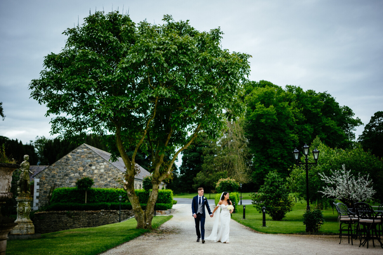 Tara and Darren’s Unforgettable Wedding at Bellingham Castle 106 A couple dressed in wedding attire walk hand in hand down a gravel path at their wedding at Bellingham Castle, surrounded by greenery, trees, and stone buildings on a cloudy day.