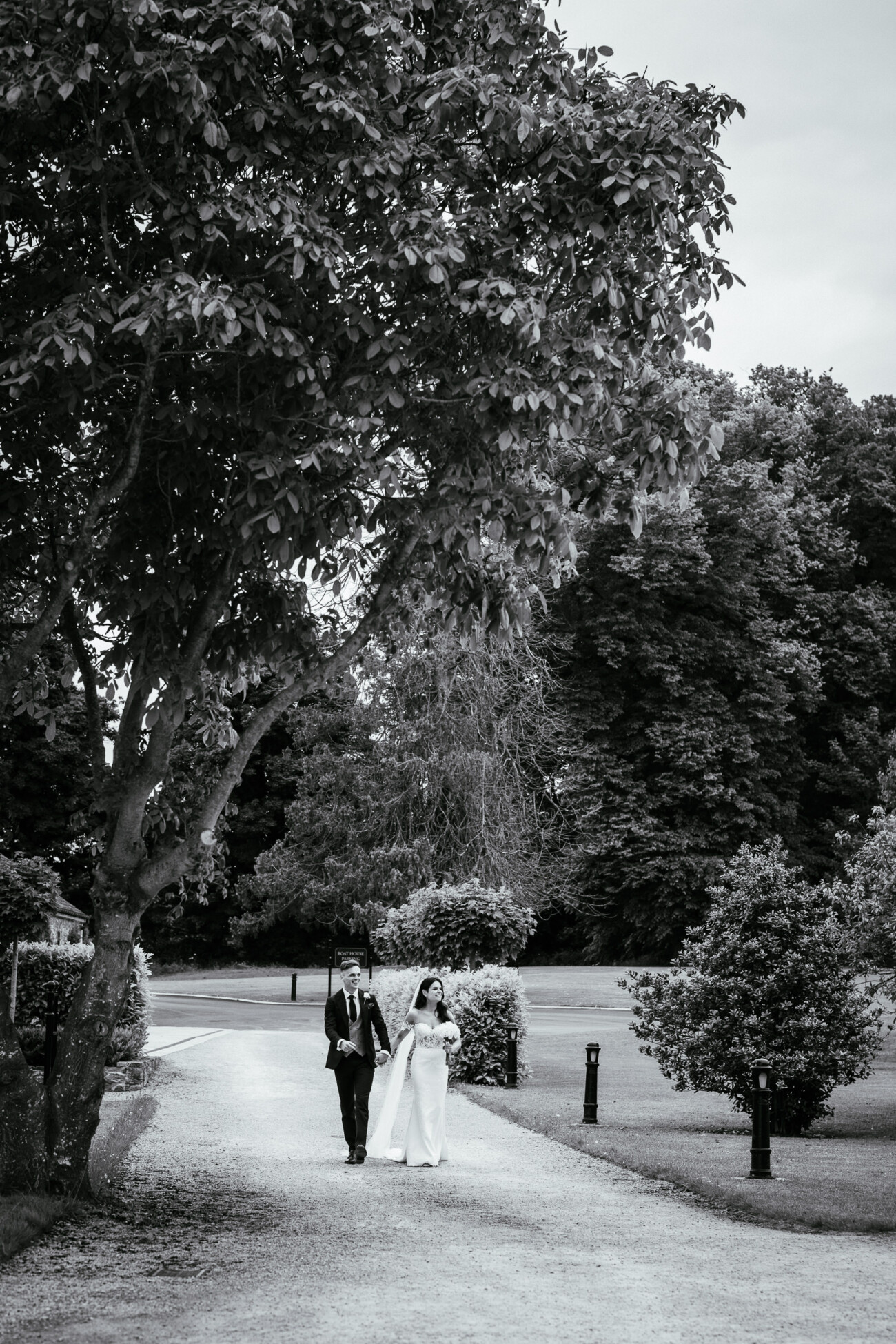 Tara and Darren’s Unforgettable Wedding at Bellingham Castle 107 A bride and groom walk together down a tree-lined path in a park or garden, dressed in wedding attire. The scene is in black and white, beautifully capturing the timeless elegance of their wedding at Bellingham Castle.