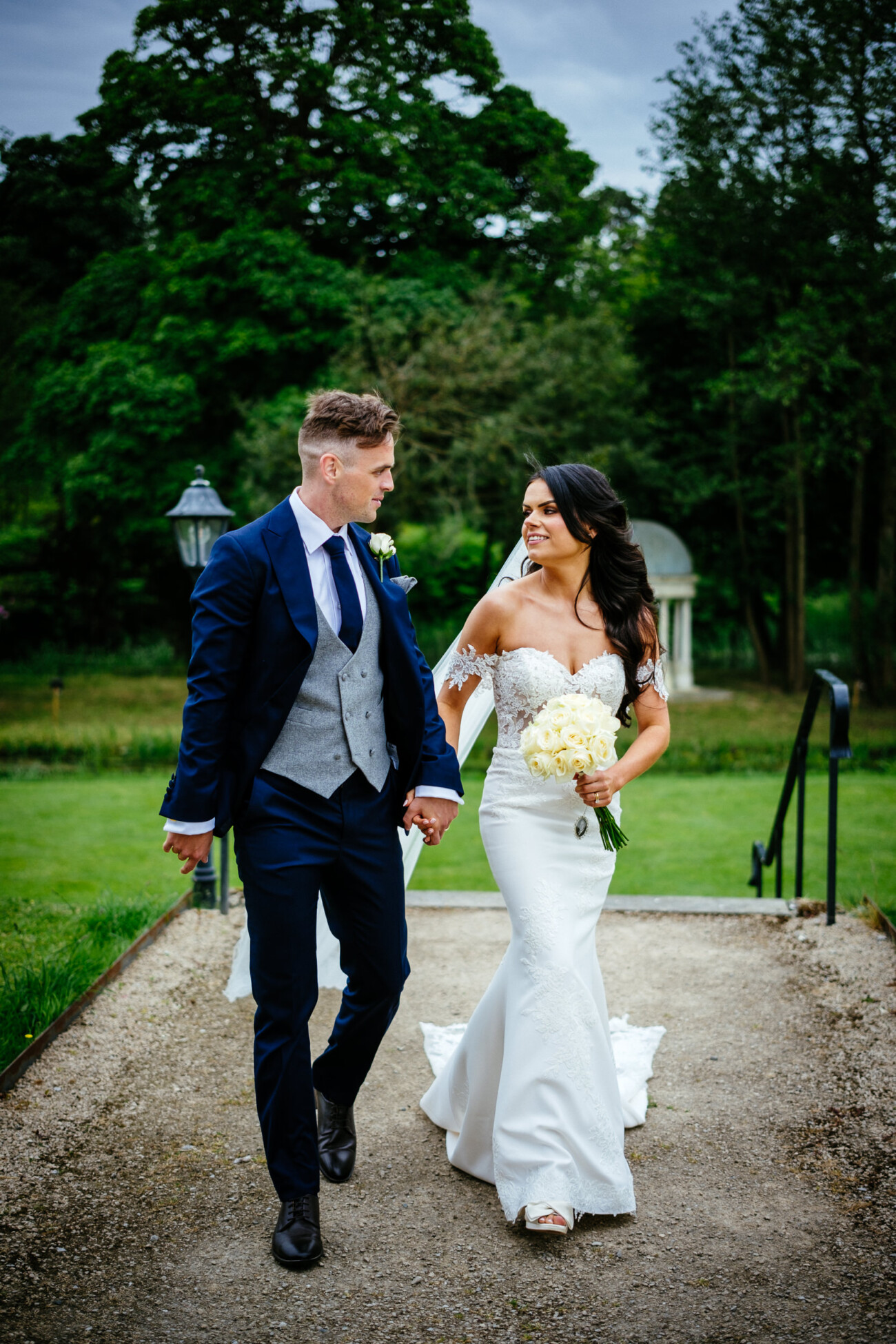 Tara and Darren’s Unforgettable Wedding at Bellingham Castle 108 A bride in a white dress and groom in a suit hold hands and walk outdoors on a path, surrounded by greenery and trees, capturing the charm of a wedding at Bellingham Castle.