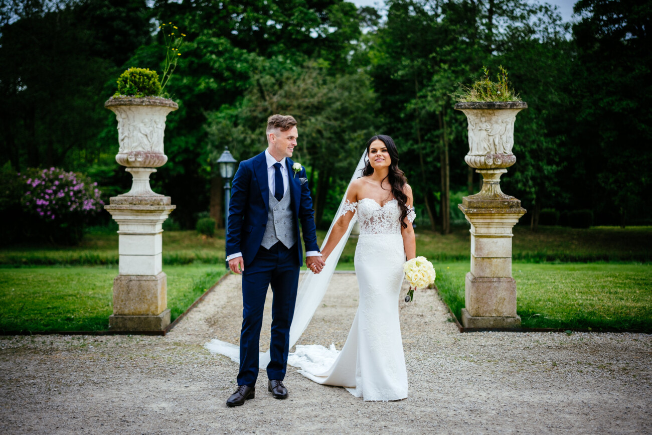 Tara and Darren’s Unforgettable Wedding at Bellingham Castle 109 A bride and groom stand outdoors, holding hands on a gravel path between two stone urns, surrounded by greenery during their wedding at Bellingham Castle.