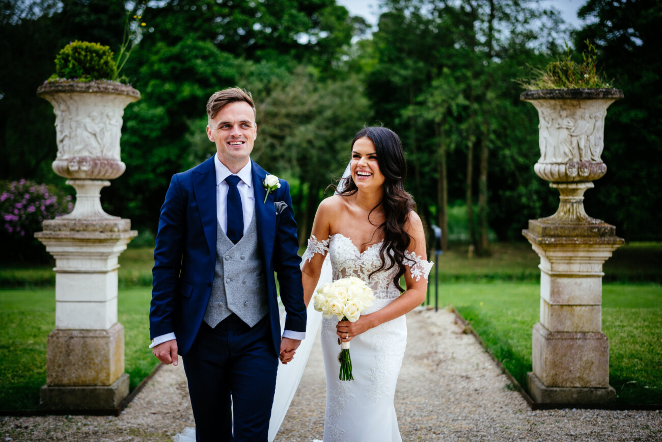 Tara and Darren’s Unforgettable Wedding at Bellingham Castle 110 A bride and groom walk outdoors, holding hands and smiling, dressed in formal wedding attire with greenery and large decorative planters in the background during their elegant wedding at Bellingham Castle.