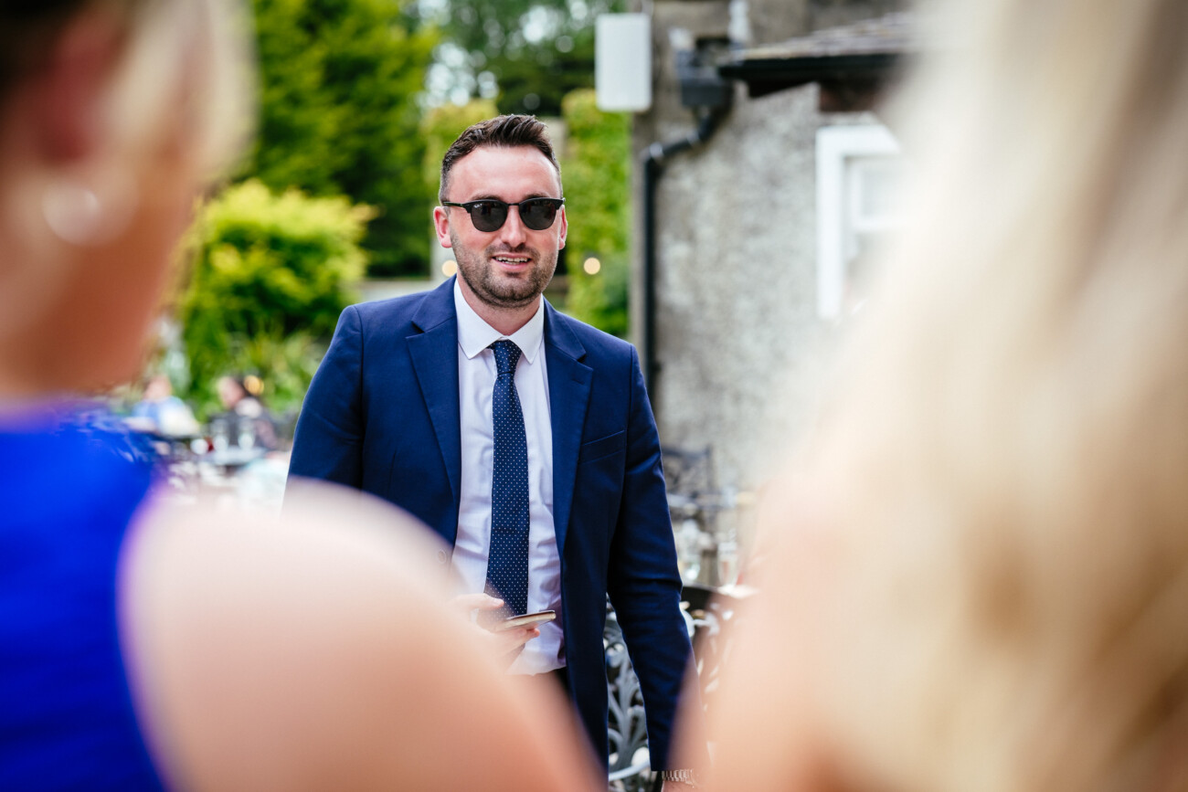 Tara and Darren’s Unforgettable Wedding at Bellingham Castle 111 Man in a blue suit and sunglasses stands outdoors, holding a mobile, facing two people with blurred blonde hair in the foreground during a wedding at Bellingham Castle.