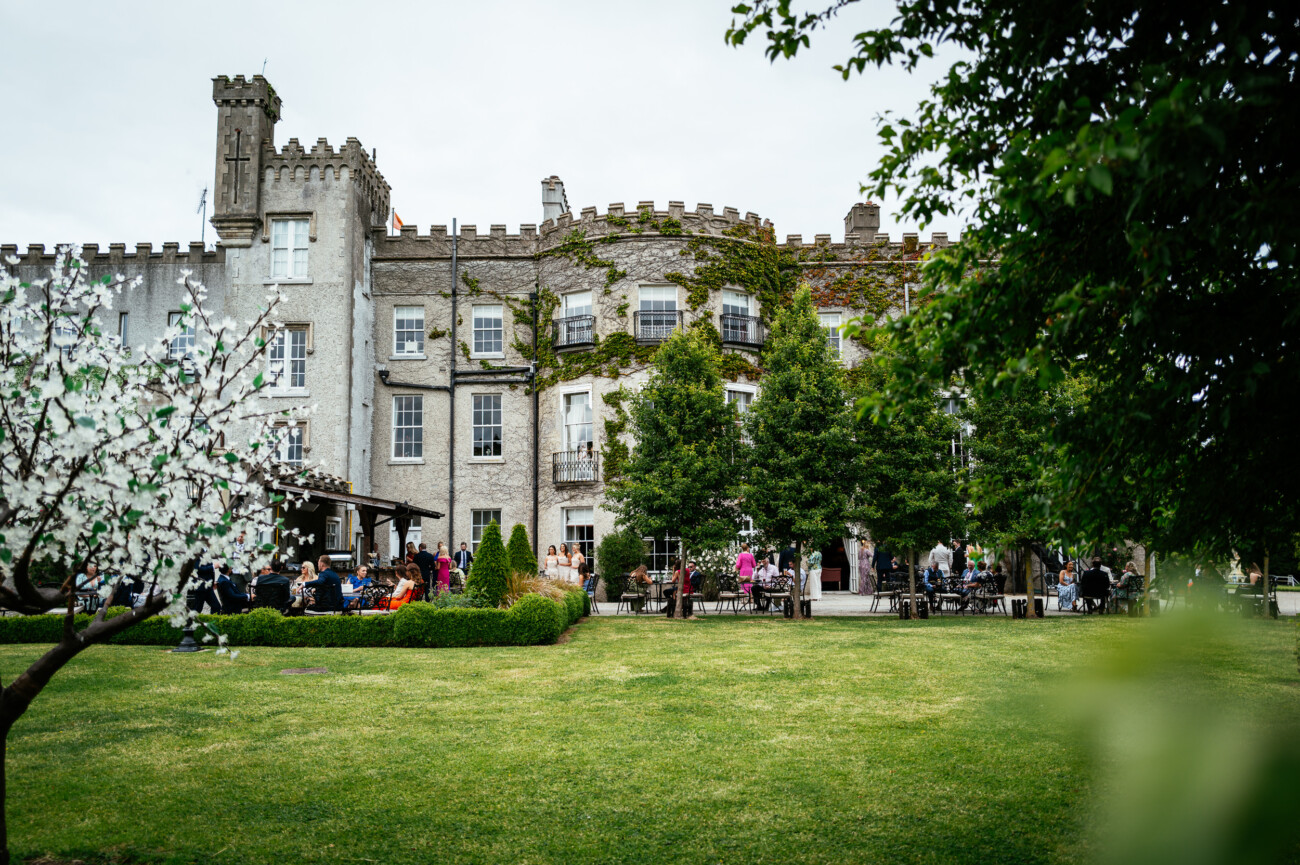 Tara and Darren’s Unforgettable Wedding at Bellingham Castle 112 A large stone castle with battlements is seen behind a garden lawn, where people are seated at outdoor tables among trees and landscaping, enjoying a beautiful wedding at Bellingham Castle.