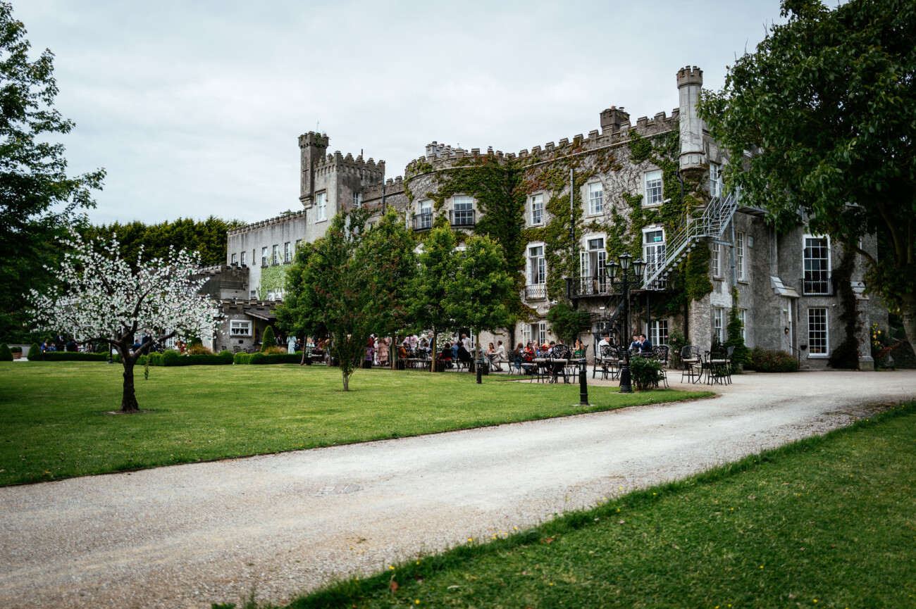Tara and Darren’s Unforgettable Wedding at Bellingham Castle 113 A stone castle covered in ivy sits behind a lawn and gravel path, with several people gathered at outdoor tables under trees, creating a picturesque setting for a wedding at Bellingham Castle.