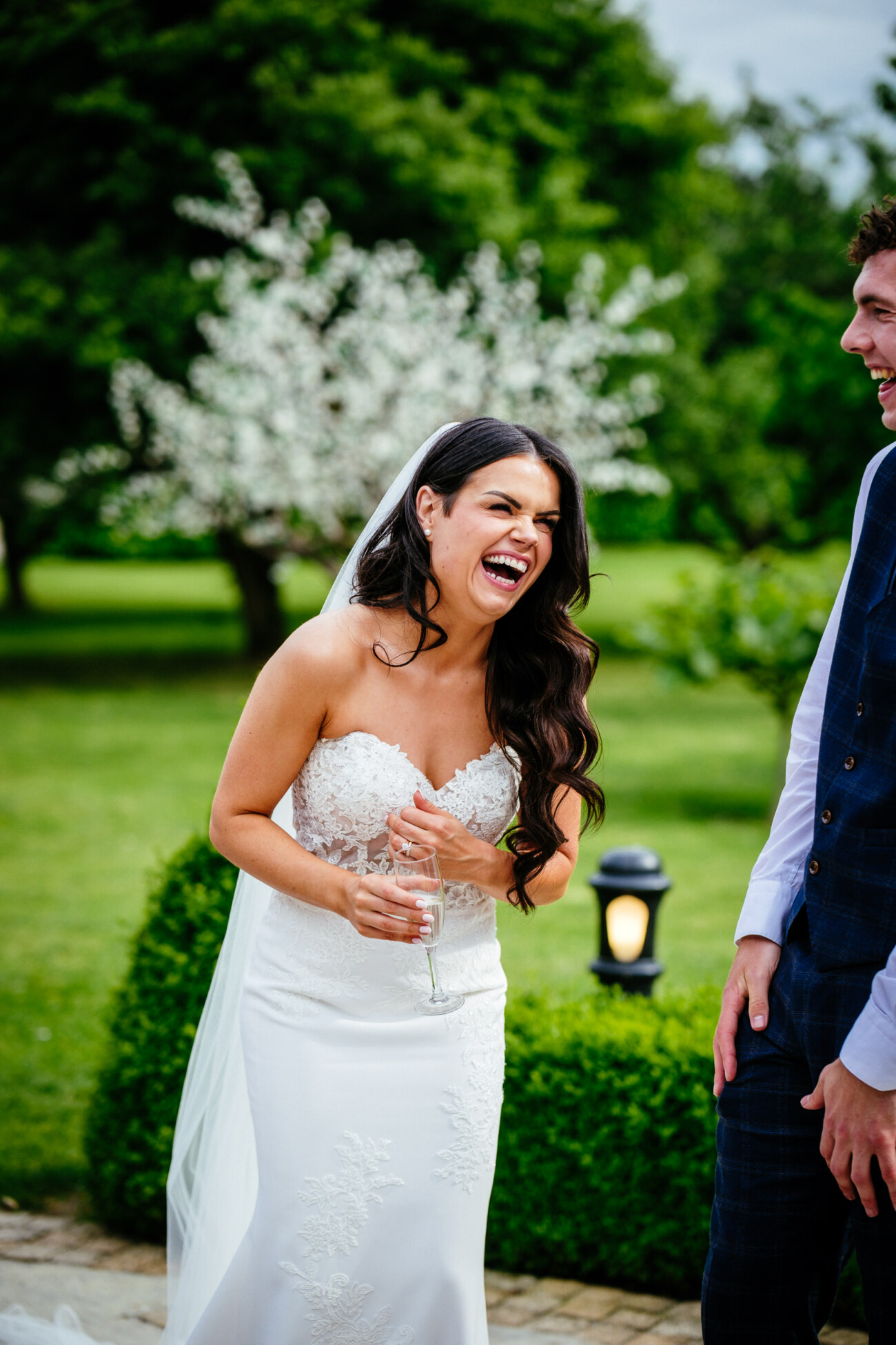 Tara and Darren’s Unforgettable Wedding at Bellingham Castle 114 A bride in a white dress and veil laughs joyfully outdoors whilst holding a glass, standing next to a person in a suit, celebrating their wedding at Bellingham Castle.