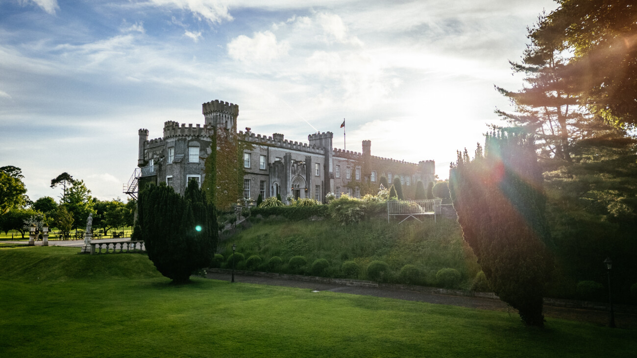 Tara and Darren’s Unforgettable Wedding at Bellingham Castle 152 Historic stone castle with crenellated towers sits atop a manicured grassy hill, surrounded by trees under a partly cloudy sky with sunlight streaming through—the perfect setting for a romantic wedding at Bellingham Castle.
