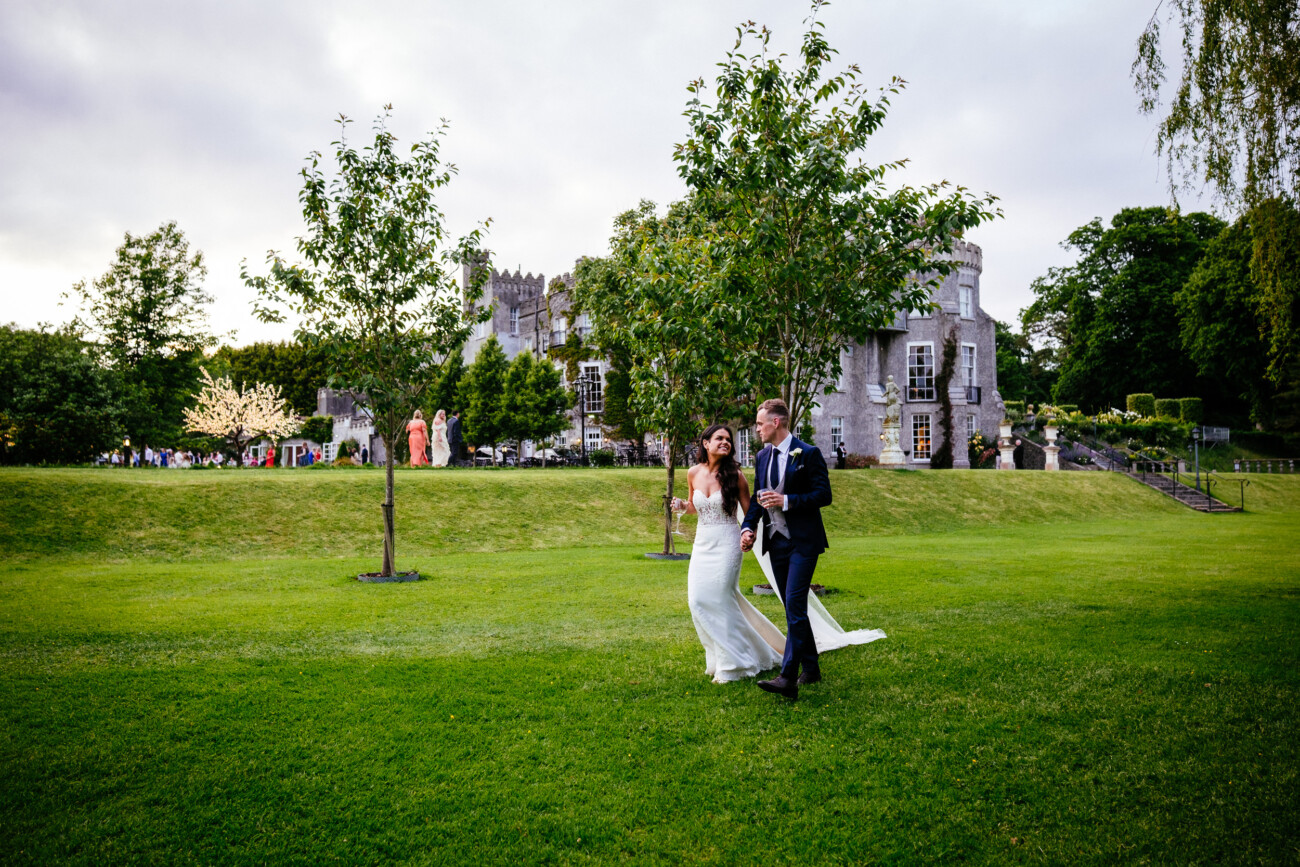 Tara and Darren’s Unforgettable Wedding at Bellingham Castle 153 A bride and groom walk across a grassy lawn near a castle during their wedding at Bellingham Castle, with trees and several people visible in the background under a cloudy sky.