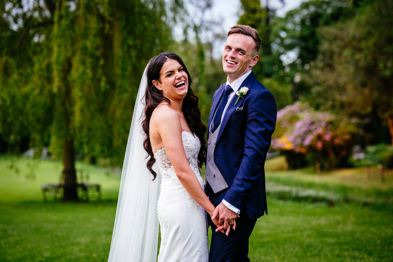 Tara and Darren’s Unforgettable Wedding at Bellingham Castle 154 The bride and groom laugh together in the gardens at Bellingham Castle, with a weeping willow and blossoming rhododendrons in the background.