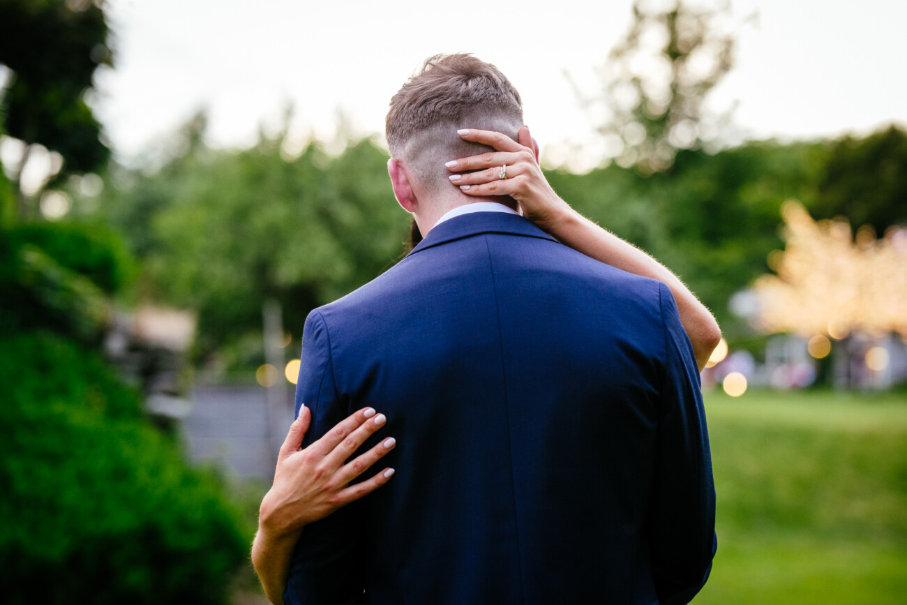 Tara and Darren’s Unforgettable Wedding at Bellingham Castle 155 A person in a blue suit stands outdoors with another person's arms wrapped around their neck and back, green trees in the background, capturing a tender moment during a wedding at Bellingham Castle.