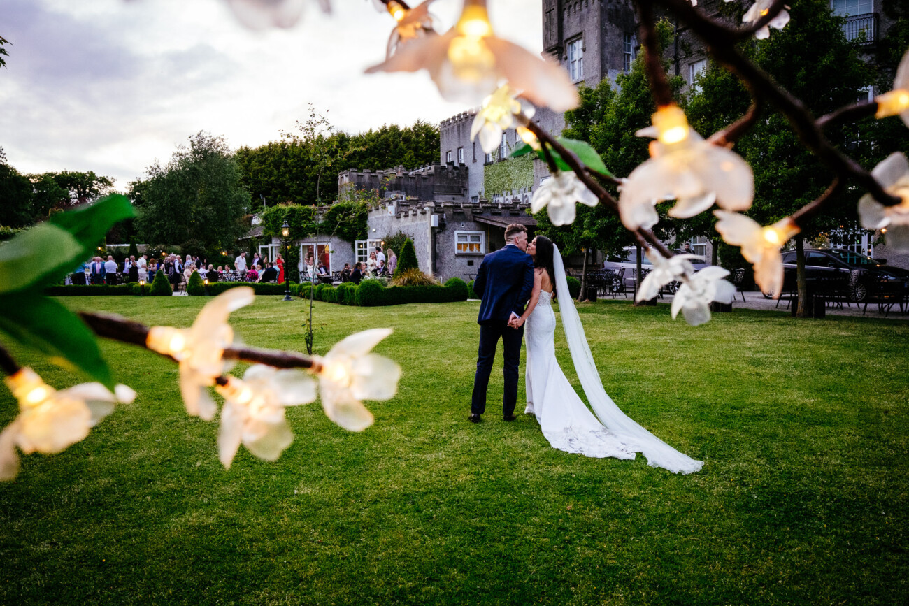 Tara and Darren’s Unforgettable Wedding at Bellingham Castle 156 A bride and groom kiss on a lawn outside Bellingham Castle, framed by white flower lights in the foreground, with wedding guests gathered in the background—capturing a magical moment at their wedding at Bellingham Castle.