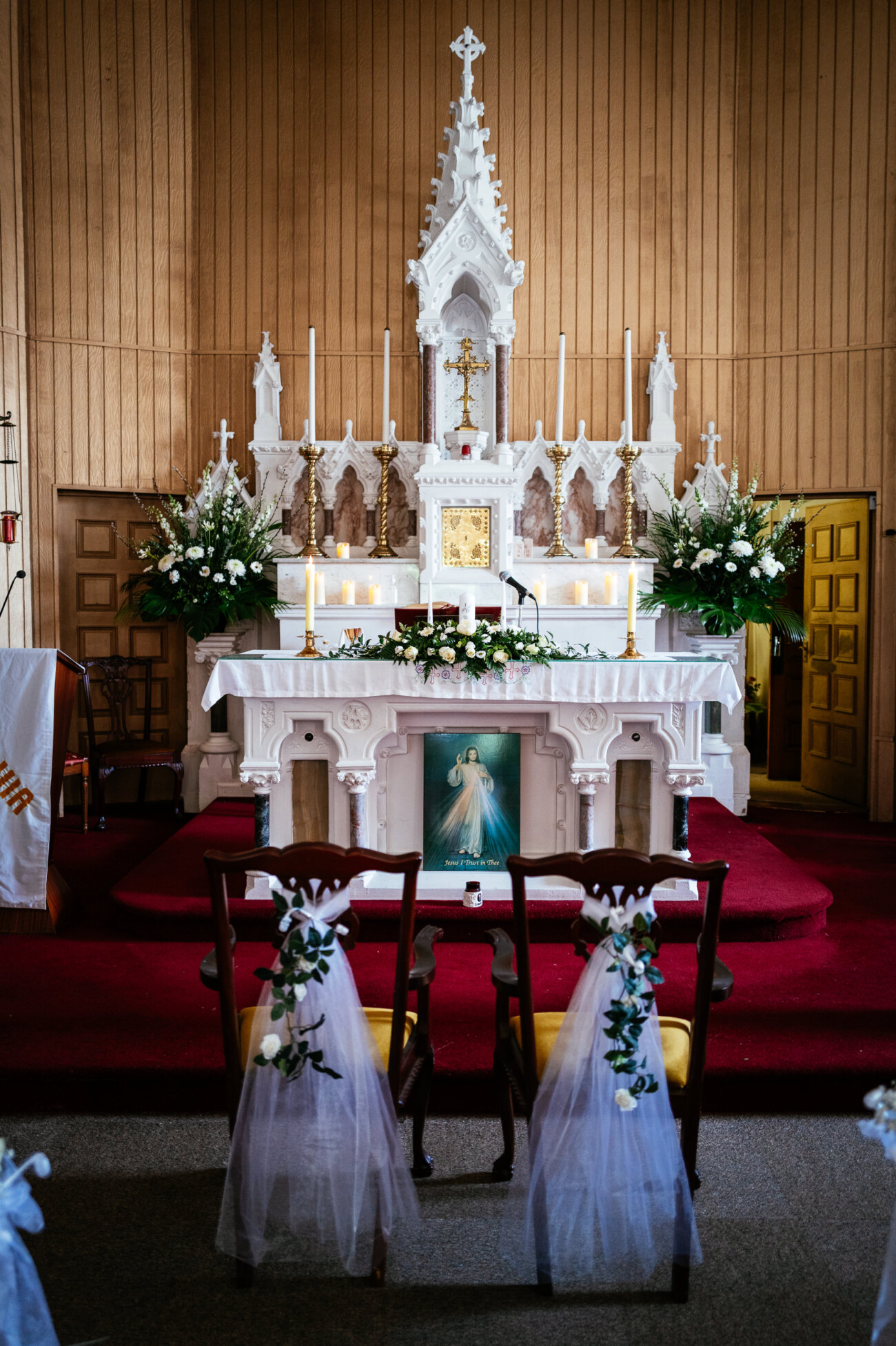 Tara and Darren’s Unforgettable Wedding at Bellingham Castle 45 A decorated church altar with white flowers, candles, and two chairs draped with white tulle and greenery in the foreground creates a romantic setting reminiscent of a wedding at Bellingham Castle.
