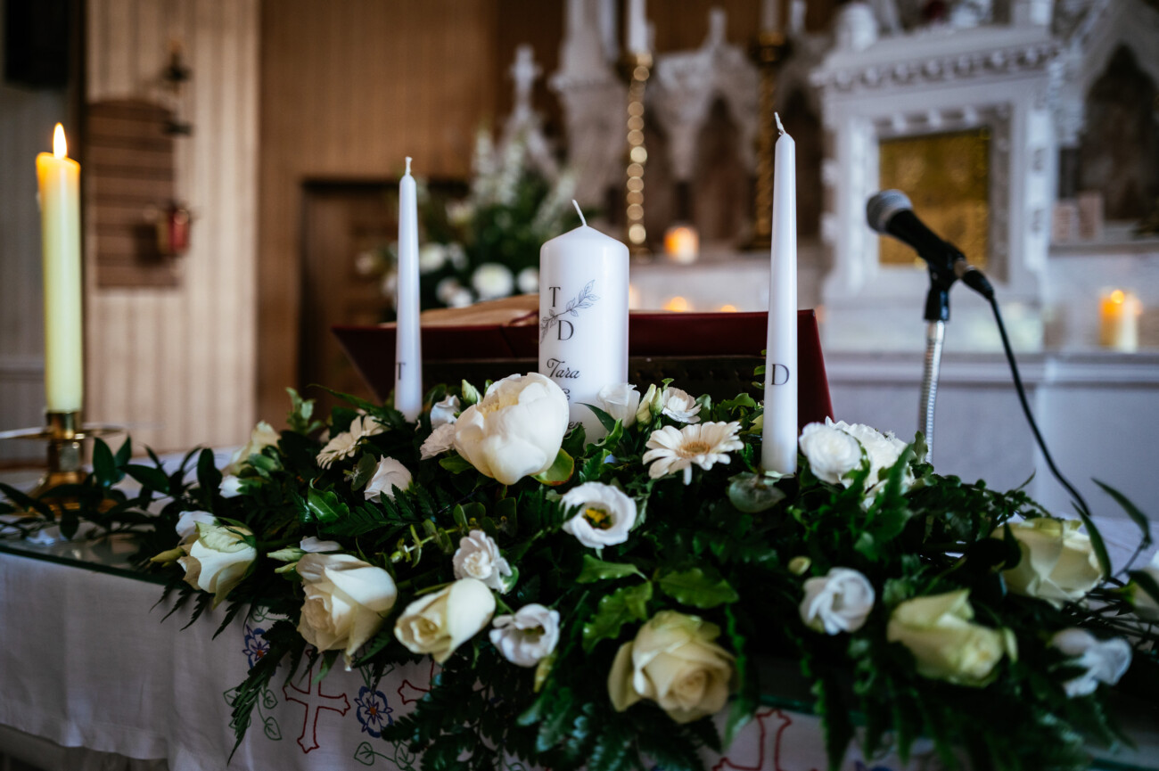 Tara and Darren’s Unforgettable Wedding at Bellingham Castle 46 A church altar decorated with white candles, white flowers, and greenery, with a microphone and a book placed on the table in front of a white and gold altar background, creating an elegant setting reminiscent of a wedding at Bellingham Castle.