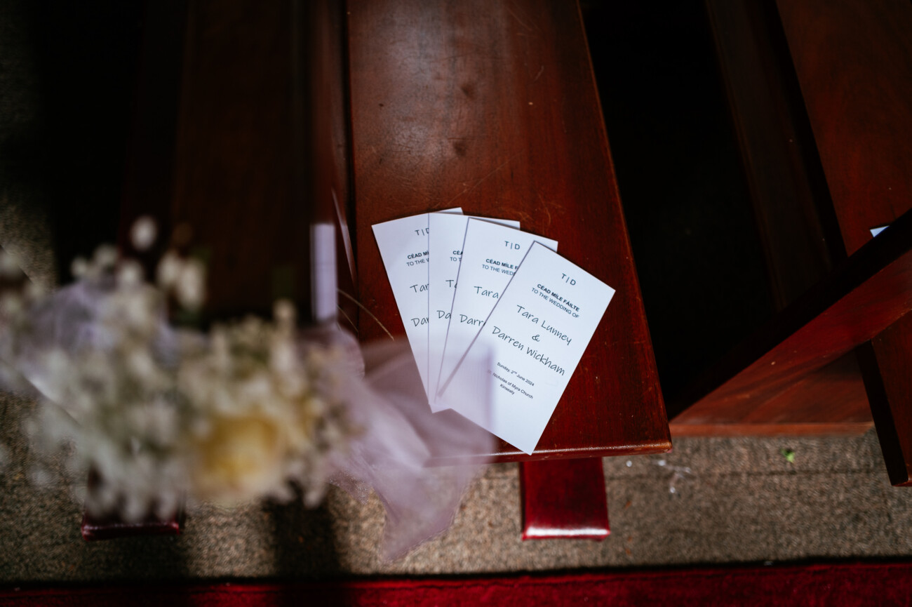 Tara and Darren’s Unforgettable Wedding at Bellingham Castle 47 Three wedding programmes are placed on a wooden pew next to a blurred bouquet of flowers, with light shining on the scene, capturing the elegant atmosphere of a wedding at Bellingham Castle.