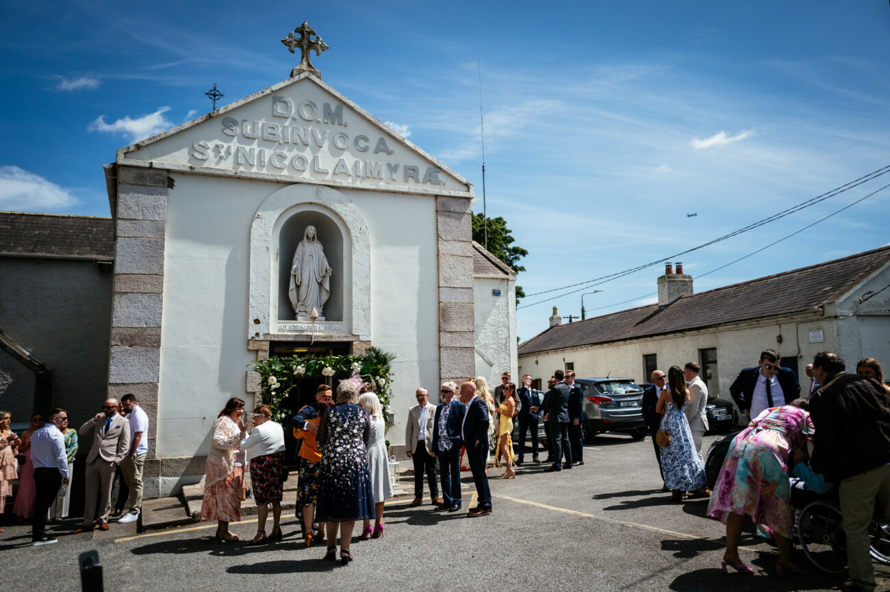 Tara and Darren’s Unforgettable Wedding at Bellingham Castle 48 A group of people gather outside a small church on a sunny day, standing and talking near parked cars and a statue of a saint above the entrance, capturing the joyful spirit reminiscent of a wedding at Bellingham Castle.