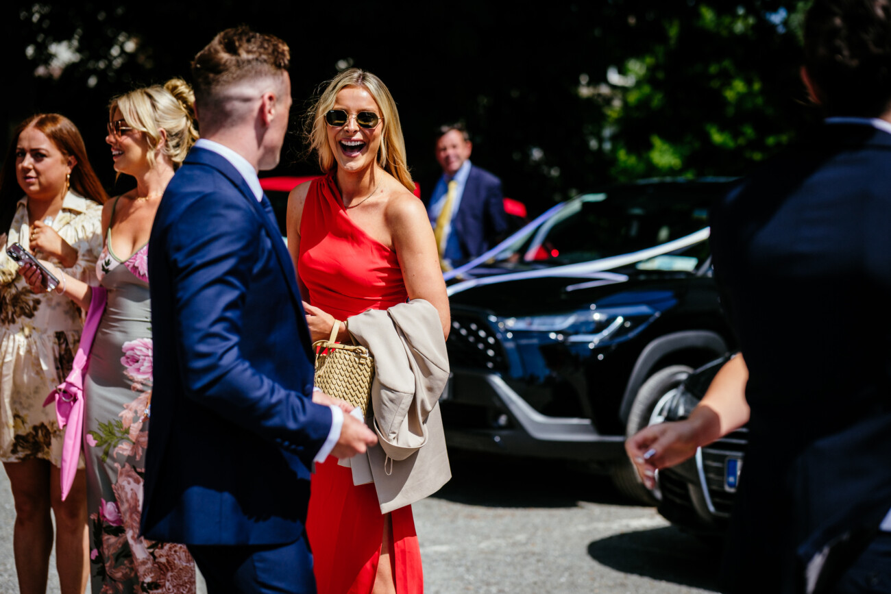 Tara and Darren’s Unforgettable Wedding at Bellingham Castle 49 A group of well-dressed people stand and talk outside near parked cars on a sunny day, enjoying the atmosphere of a wedding at Bellingham Castle. One woman in a red dress is smiling and holding a beige coat and woven handbag.