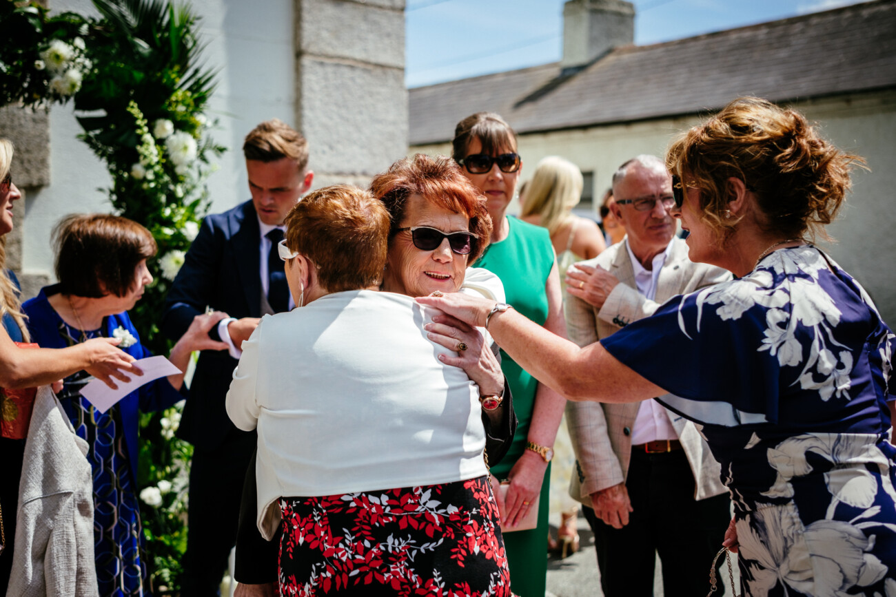 Tara and Darren’s Unforgettable Wedding at Bellingham Castle 51 A group of people interact and embrace outdoors at a social event, with women in the foreground and stone buildings in the background, capturing the joyful atmosphere of a wedding at Bellingham Castle.