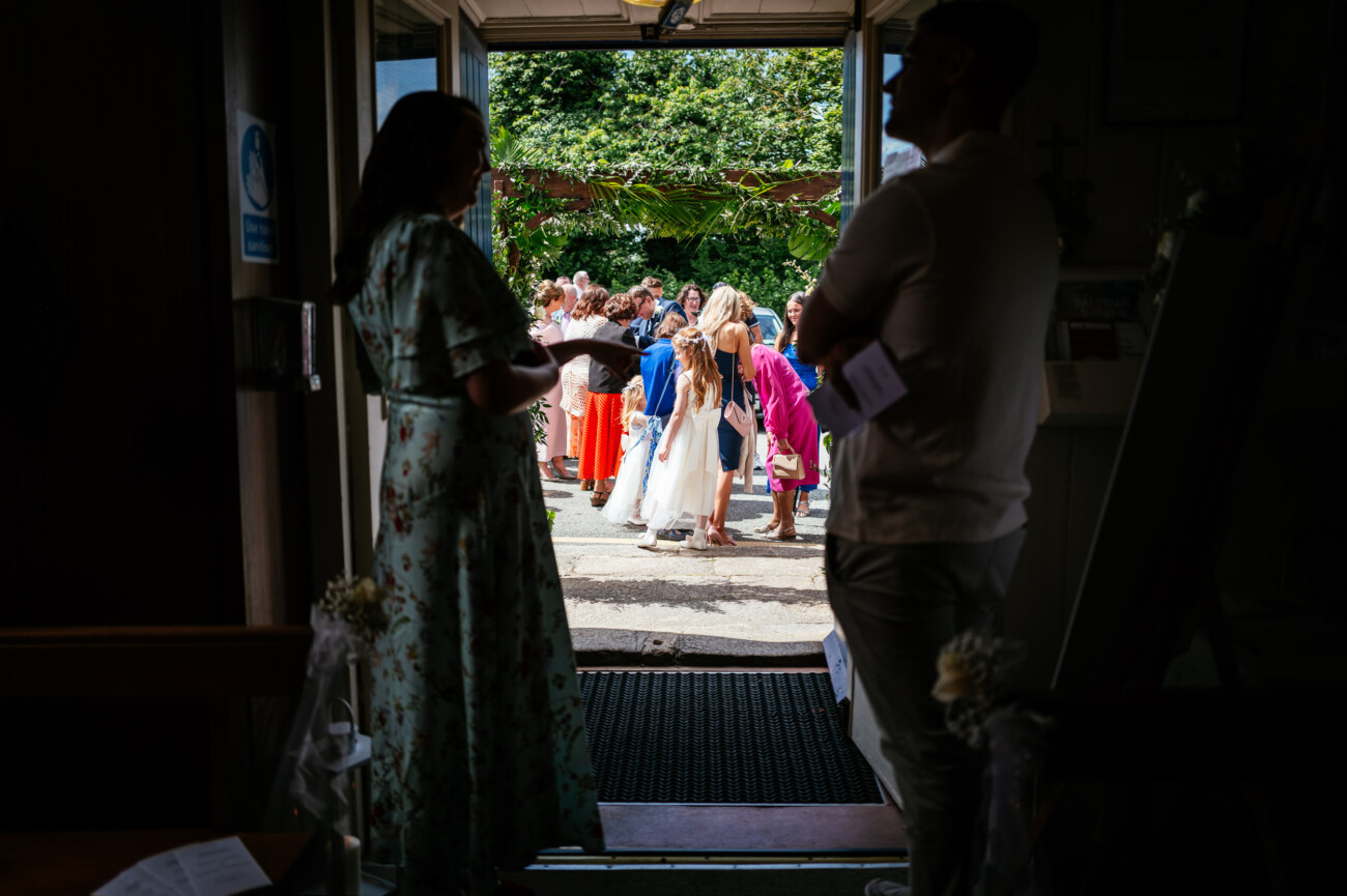 Tara and Darren’s Unforgettable Wedding at Bellingham Castle 53 People gather outside in bright daylight, viewed from a shaded doorway where two people stand in silhouette, observing the scene. Guests appear dressed for a formal or celebratory occasion, capturing the elegant atmosphere of a wedding at Bellingham Castle.