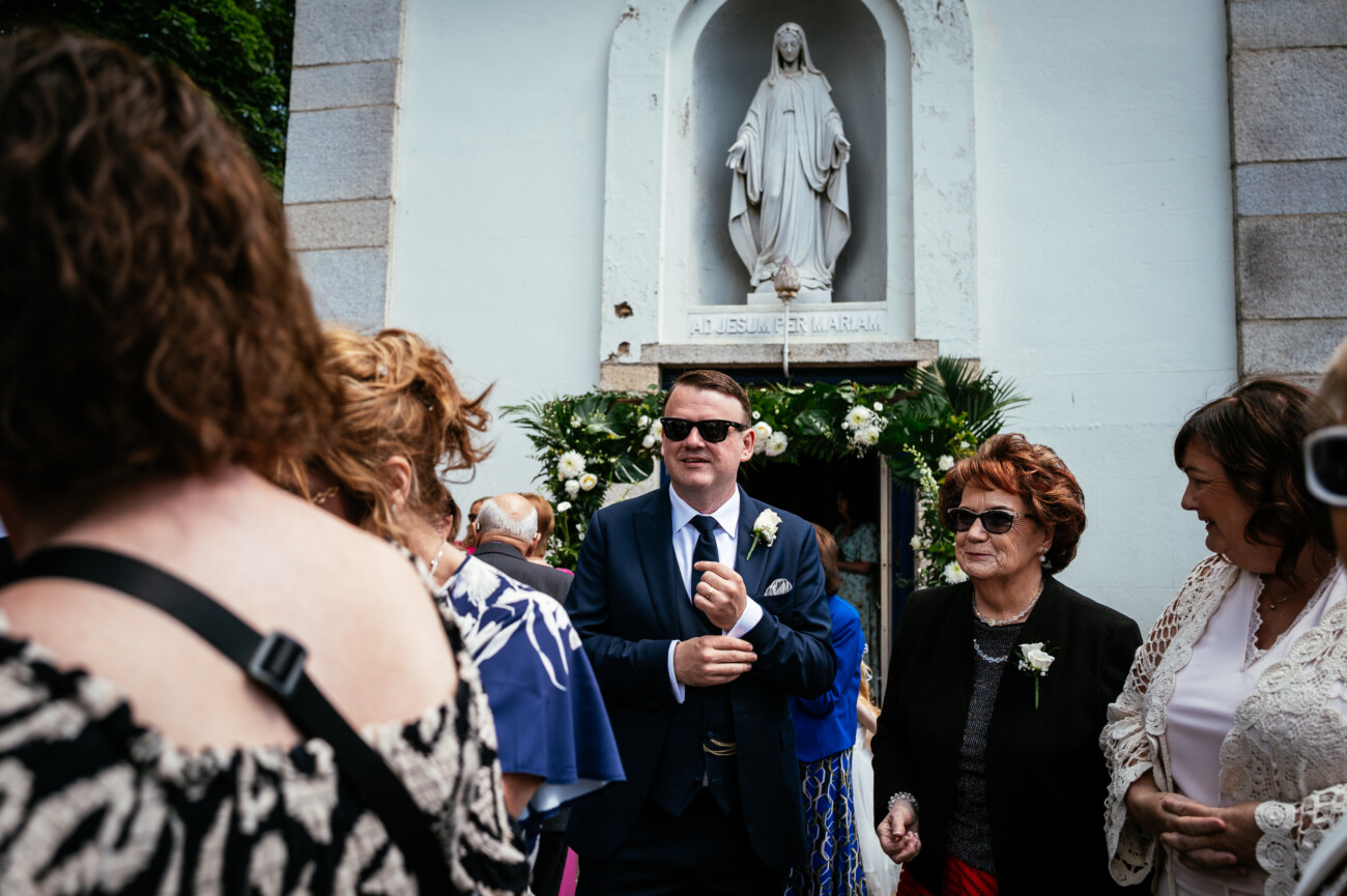 Tara and Darren’s Unforgettable Wedding at Bellingham Castle 54 People gather outside a church with a statue of Mary above the entrance, reminiscent of scenes from a wedding at Bellingham Castle. A man in a suit and sunglasses stands beside women dressed formally, surrounded by elegant floral arrangements.