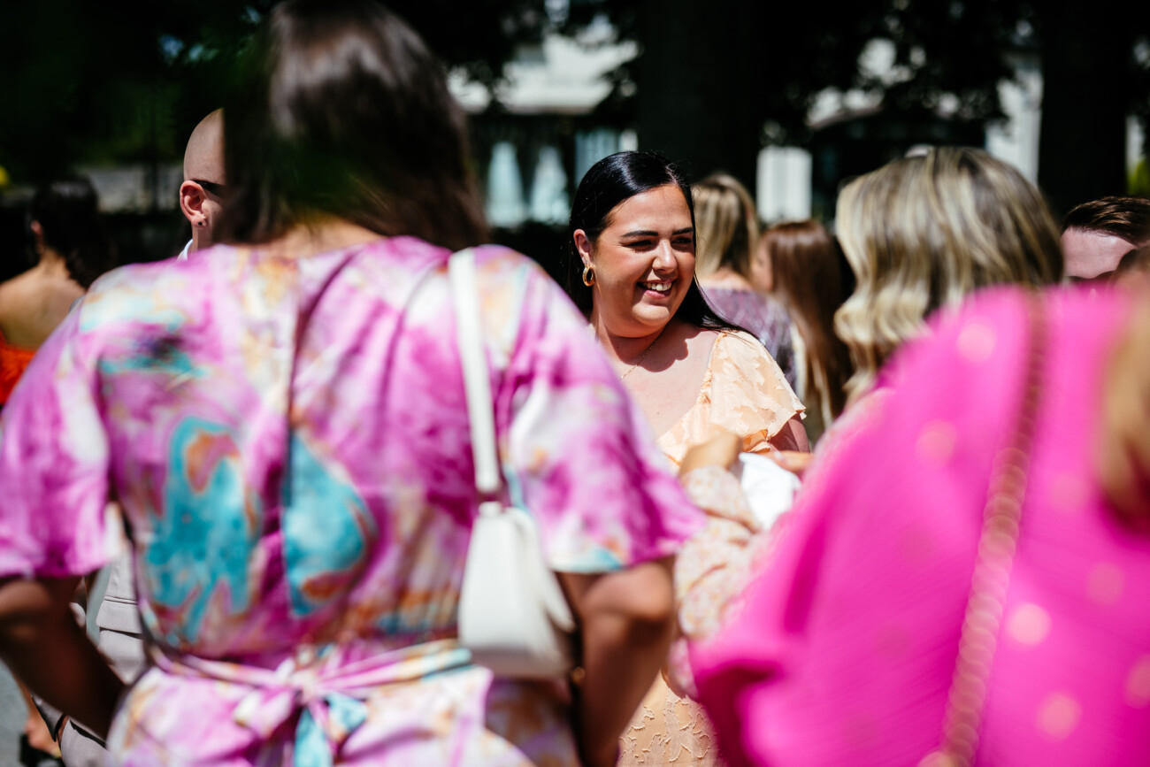 Tara and Darren’s Unforgettable Wedding at Bellingham Castle 55 A woman in a peach dress smiles whilst standing among people, some wearing vibrant pink and purple outfits, at an outdoor gathering during a wedding at Bellingham Castle.