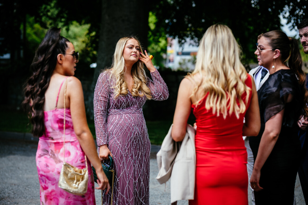 Tara and Darren’s Unforgettable Wedding at Bellingham Castle 56 Five people dressed in formal attire stand outdoors talking during a wedding at Bellingham Castle; one woman in a patterned dress touches her hair whilst others face her.
