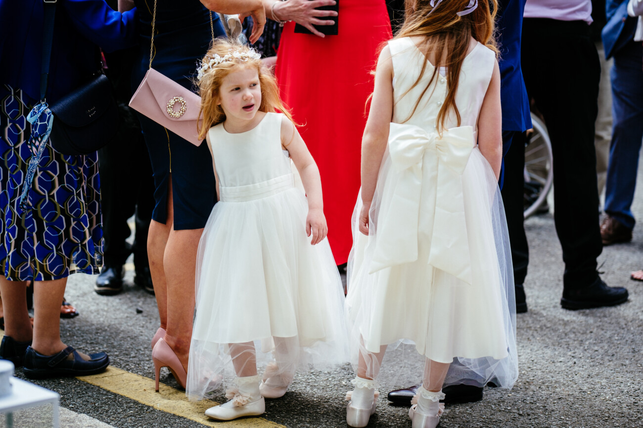 Tara and Darren’s Unforgettable Wedding at Bellingham Castle 57 Two young girls in white dresses stand among a group of adults at an outdoor event, capturing the charm of a wedding at Bellingham Castle. One girl faces forwards, the other backwards, while the adults around them are dressed in formal attire.