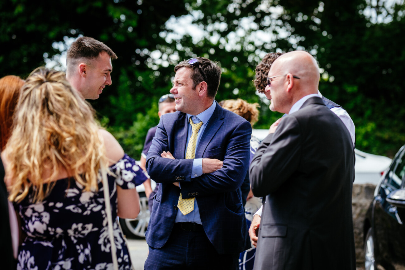 Tara and Darren’s Unforgettable Wedding at Bellingham Castle 58 A group of people in formal attire stand outdoors in conversation at a wedding at Bellingham Castle, with trees and parked cars in the background.