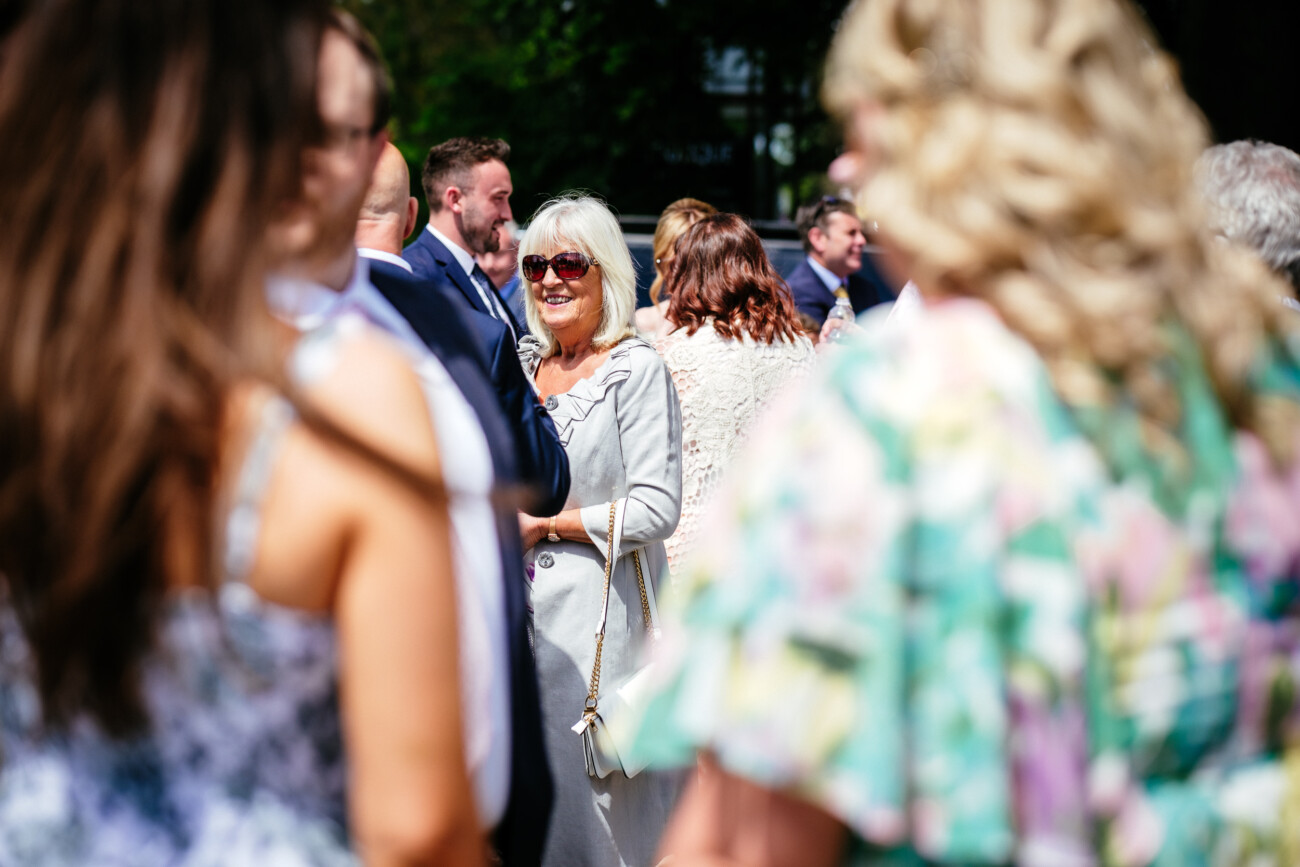 Tara and Darren’s Unforgettable Wedding at Bellingham Castle 59 A woman in sunglasses and a light outfit smiles whilst standing among a crowd of people outdoors at a sunny wedding at Bellingham Castle.