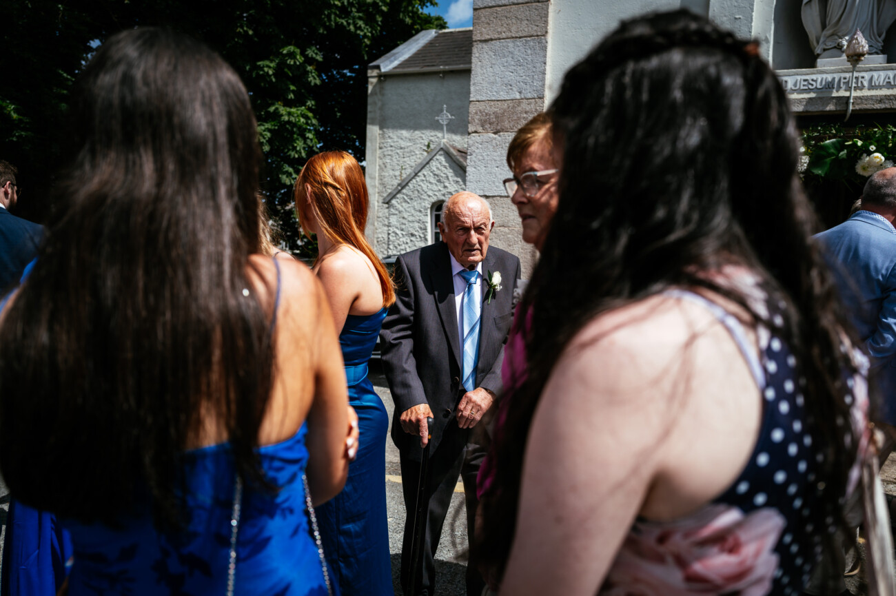 Tara and Darren’s Unforgettable Wedding at Bellingham Castle 60 An older man in a suit stands among a group of people, mostly women in dresses, outside on a sunny day near a stone building, capturing the joyful atmosphere of a wedding at Bellingham Castle.