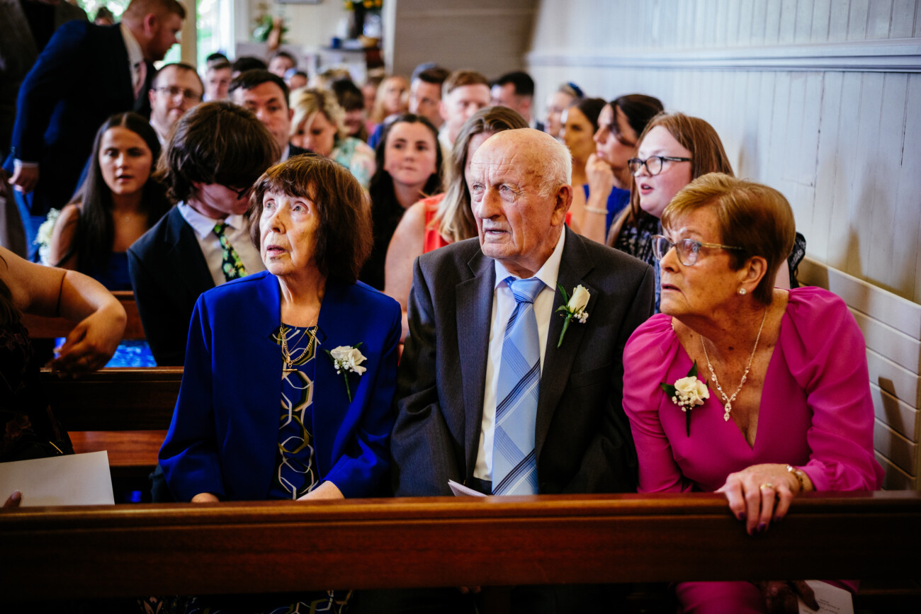 Tara and Darren’s Unforgettable Wedding at Bellingham Castle 62 Three older adults sit in the front row of a crowded church, dressed formally, with other attendees seated behind them, all gathered to celebrate a beautiful wedding at Bellingham Castle.