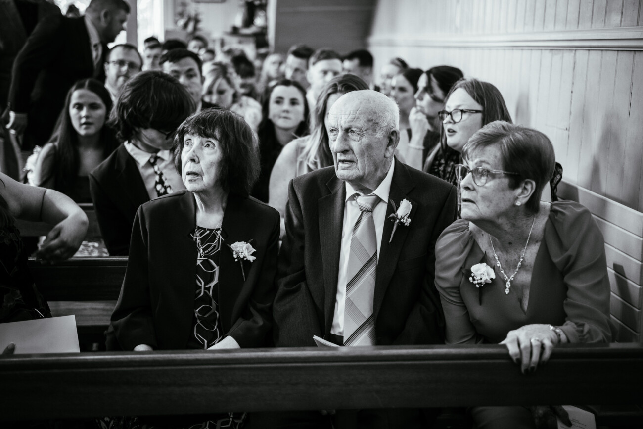 Tara and Darren’s Unforgettable Wedding at Bellingham Castle 63 Three elderly people sit in the front row of a crowded church pew, looking forward, with a group of people seated behind them at a wedding at Bellingham Castle.