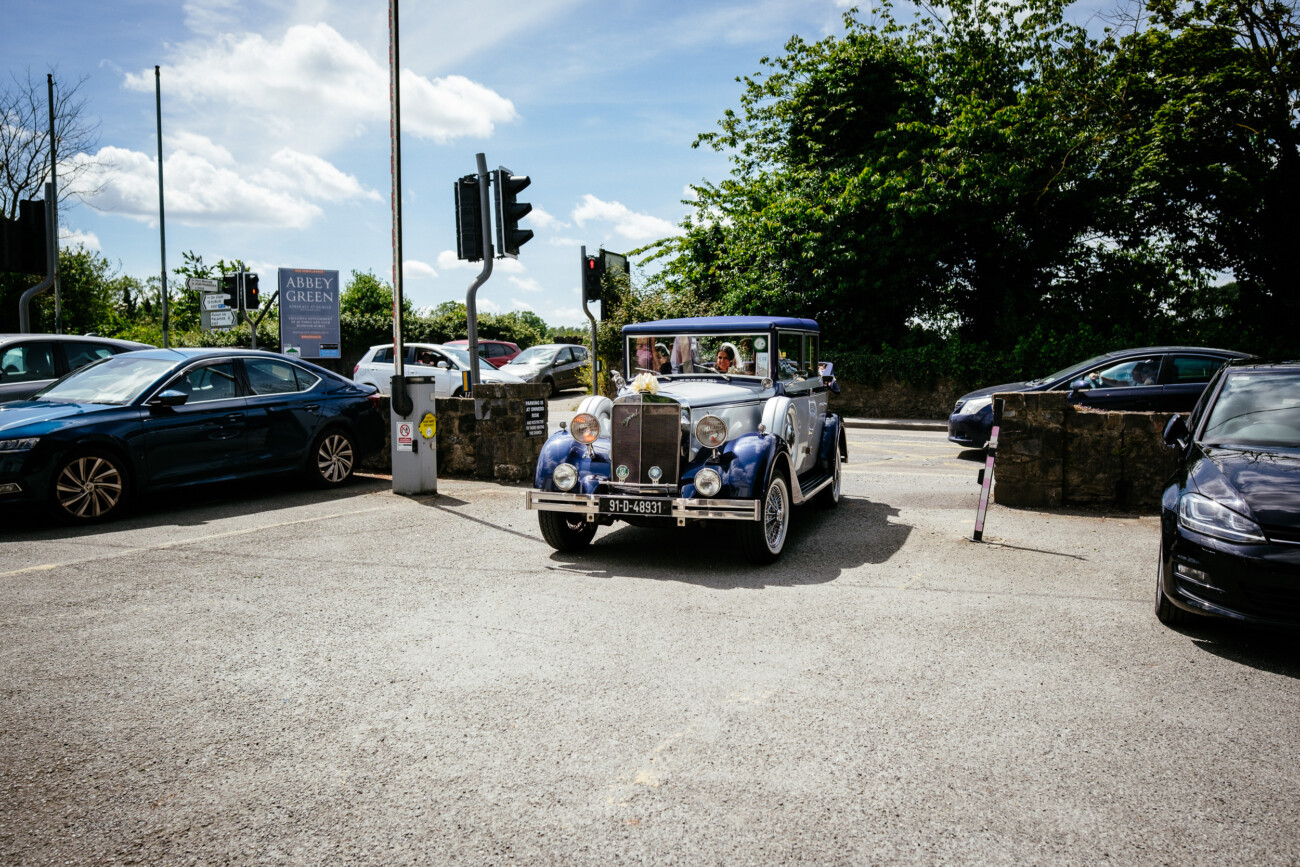 Tara and Darren’s Unforgettable Wedding at Bellingham Castle 65 A vintage blue and white car, reminiscent of those seen at a wedding at Bellingham Castle, drives through a car park surrounded by modern parked vehicles, trees, and a street with traffic lights in the background.
