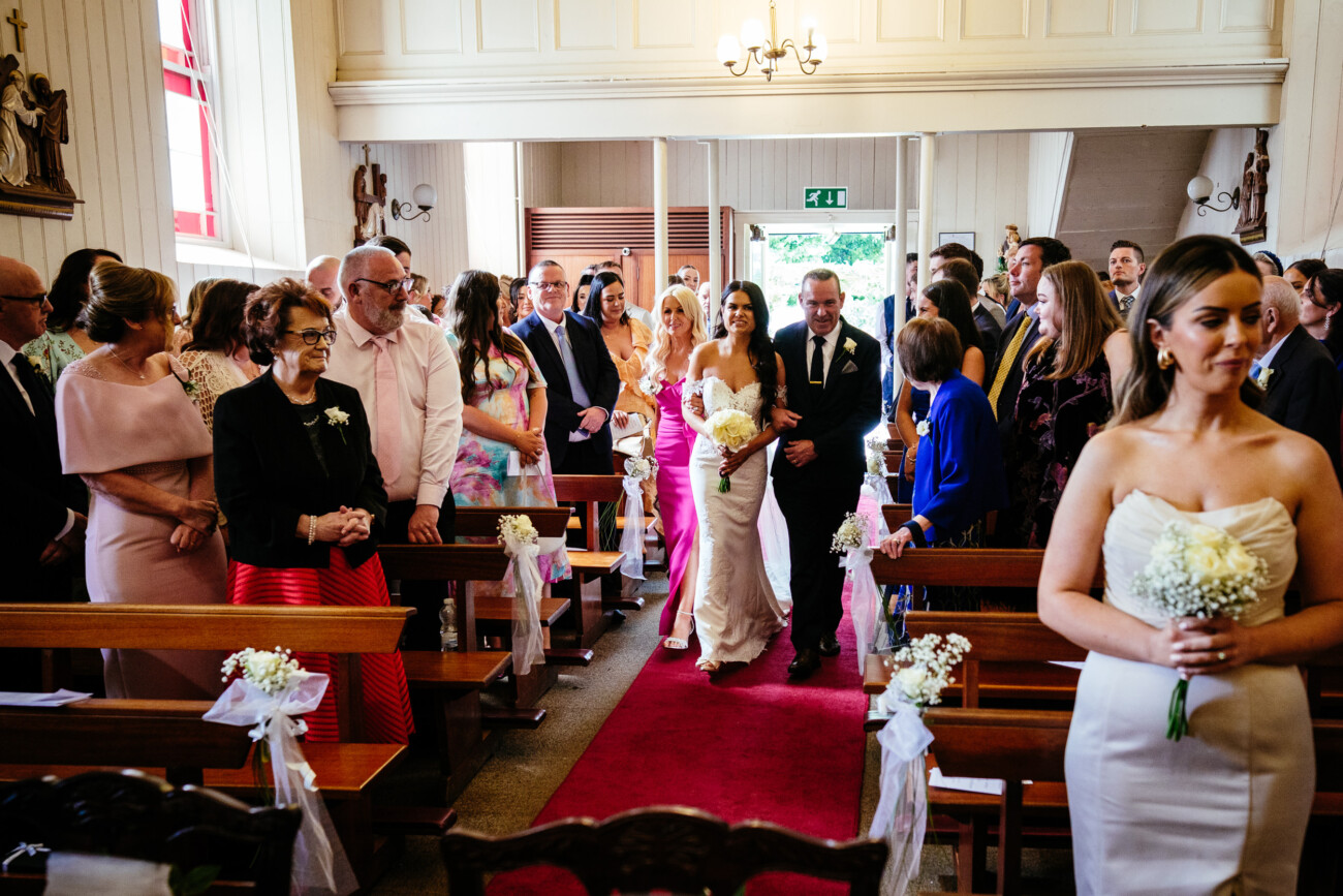 Tara and Darren’s Unforgettable Wedding at Bellingham Castle 67 A bride in a white dress walks down the aisle with an older man in a suit, as guests seated in pews watch and smile inside the church during a beautiful wedding at Bellingham Castle.
