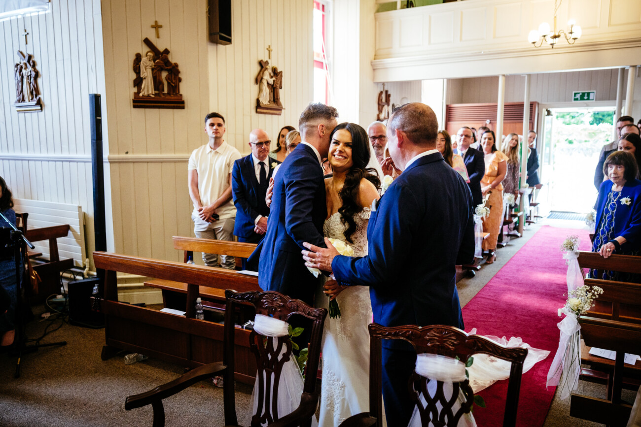 Tara and Darren’s Unforgettable Wedding at Bellingham Castle 68 A bride smiles as her father hands her to the groom at the altar in a church during their wedding at Bellingham Castle, with guests seated and standing in the background.