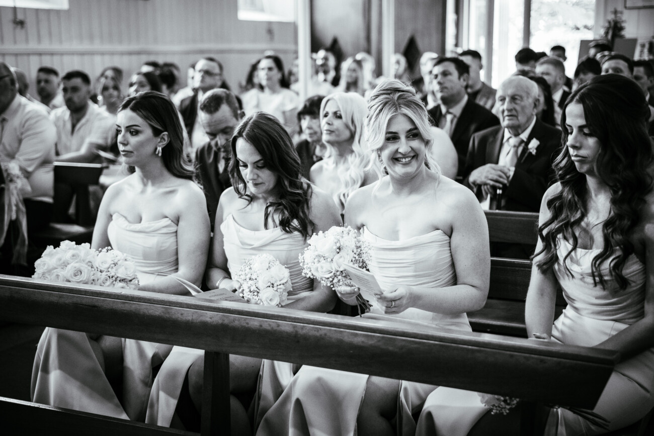 Tara and Darren’s Unforgettable Wedding at Bellingham Castle 69 Four women in matching dresses sit holding bouquets in a crowded church, surrounded by guests attending a formal event for a beautiful wedding at Bellingham Castle.