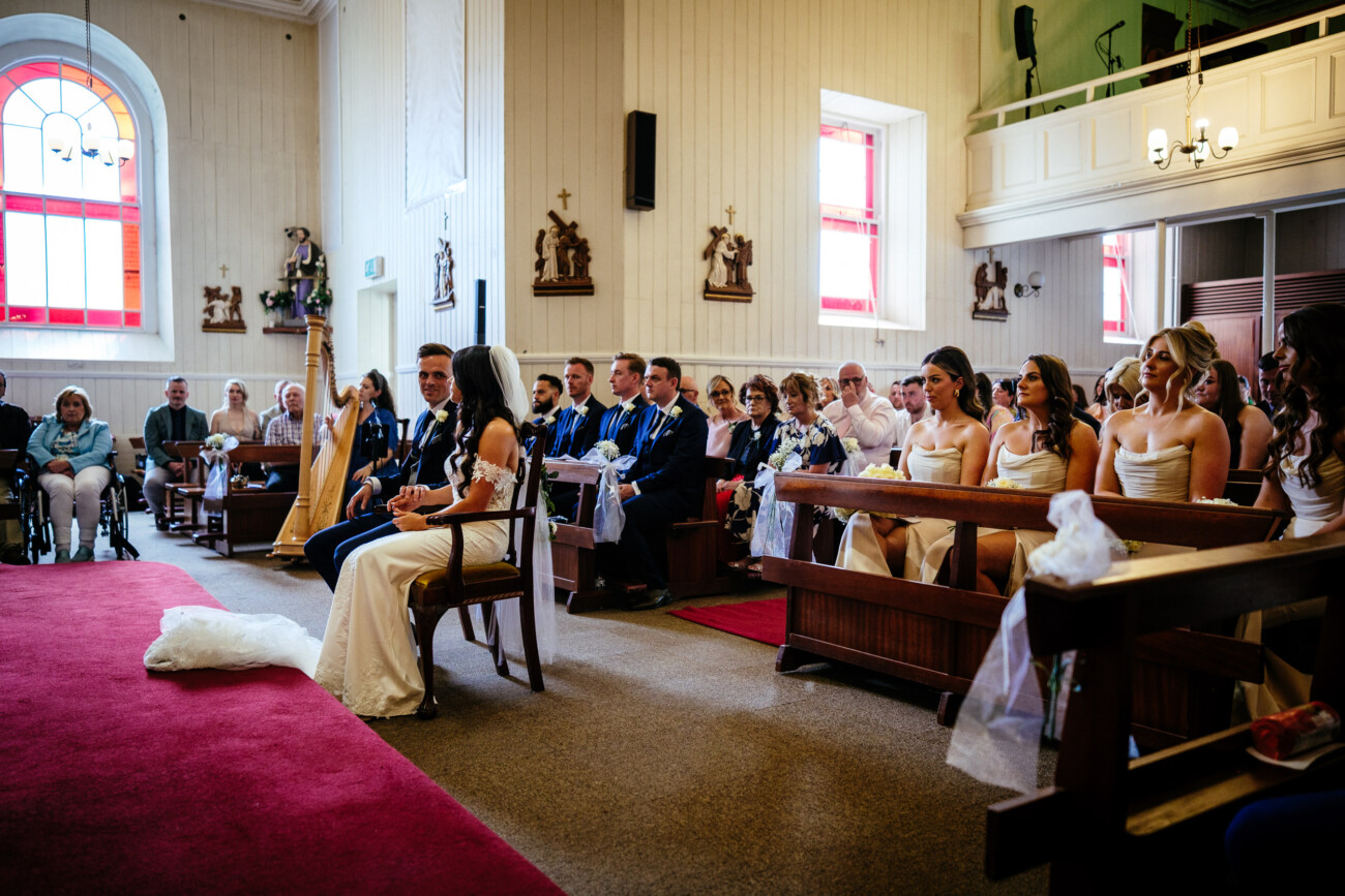 Tara and Darren’s Unforgettable Wedding at Bellingham Castle 71 Bride and groom sit at the front of a church with bridesmaids, groomsmen, and guests seated behind them during a beautiful wedding at Bellingham Castle.