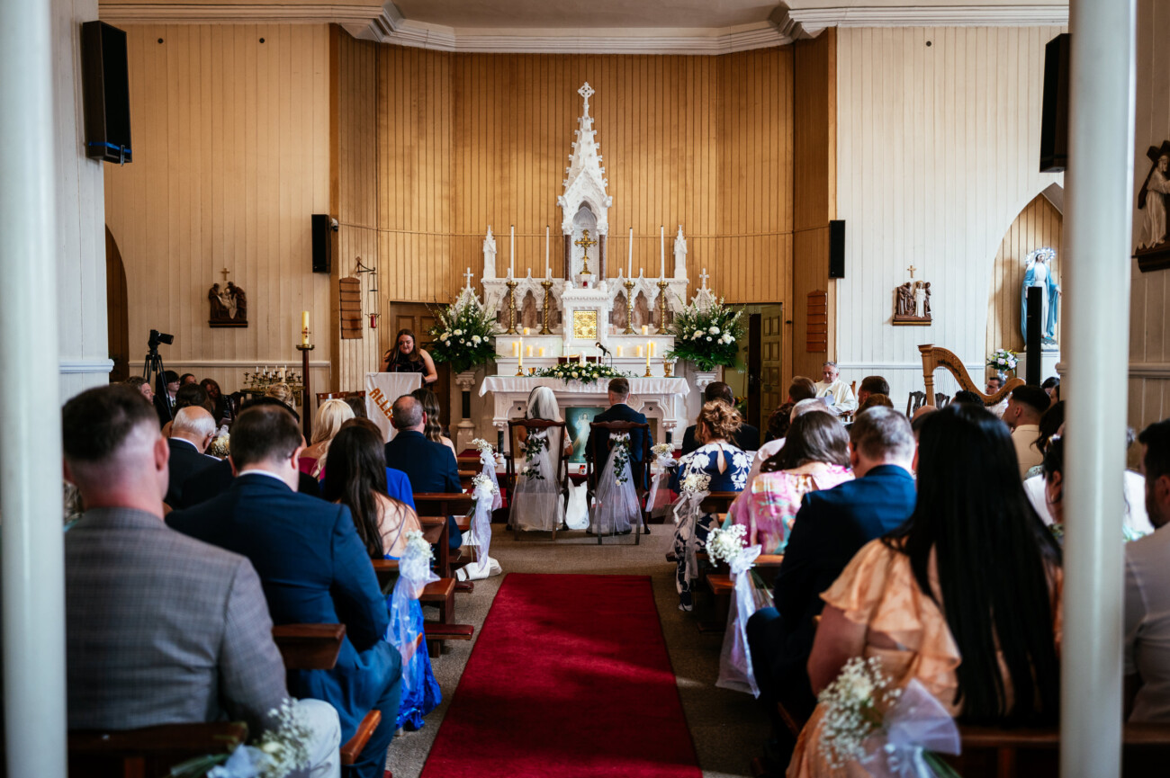 Tara and Darren’s Unforgettable Wedding at Bellingham Castle 72 A wedding at Bellingham Castle takes place in a beautifully adorned church, with guests seated in pews, facing an altar decorated with elegant flowers.