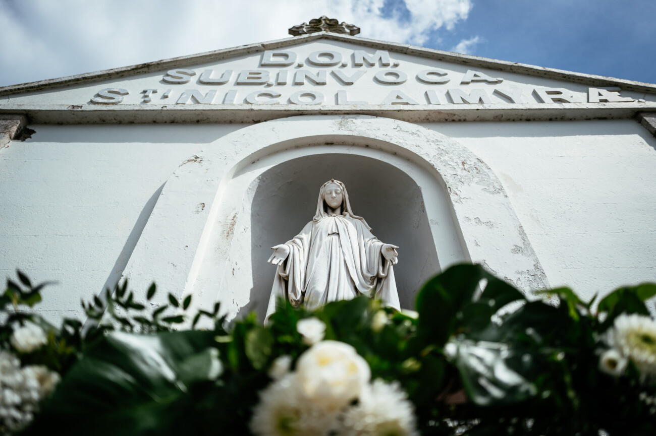 Tara and Darren’s Unforgettable Wedding at Bellingham Castle 73 A statue of the Virgin Mary stands in a niche on a white building façade with a Latin inscription, framed by white flowers and greenery in the foreground—a serene setting reminiscent of a picturesque wedding at Bellingham Castle.