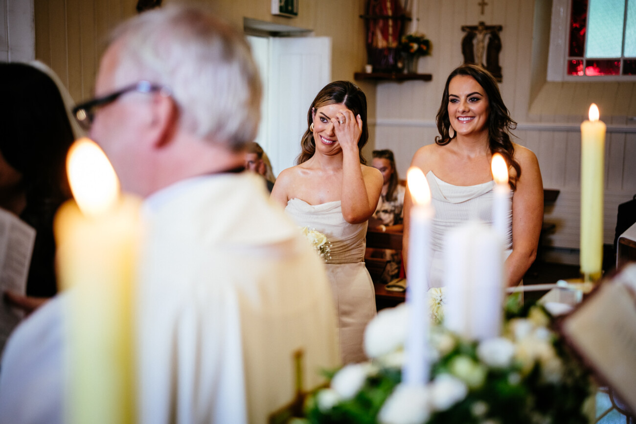 Tara and Darren’s Unforgettable Wedding at Bellingham Castle 74 Two bridesmaids in white dresses stand in a church during a wedding at Bellingham Castle, one smiling and the other wiping a tear, with candles and flowers visible in the foreground.