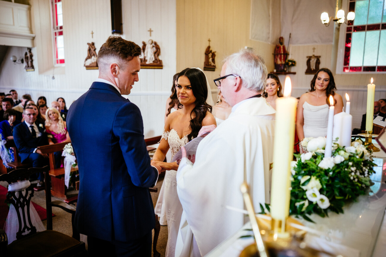 Tara and Darren’s Unforgettable Wedding at Bellingham Castle 75 Bride and groom face each other holding hands at the altar during a wedding at Bellingham Castle, with a priest officiating and guests seated in the background.