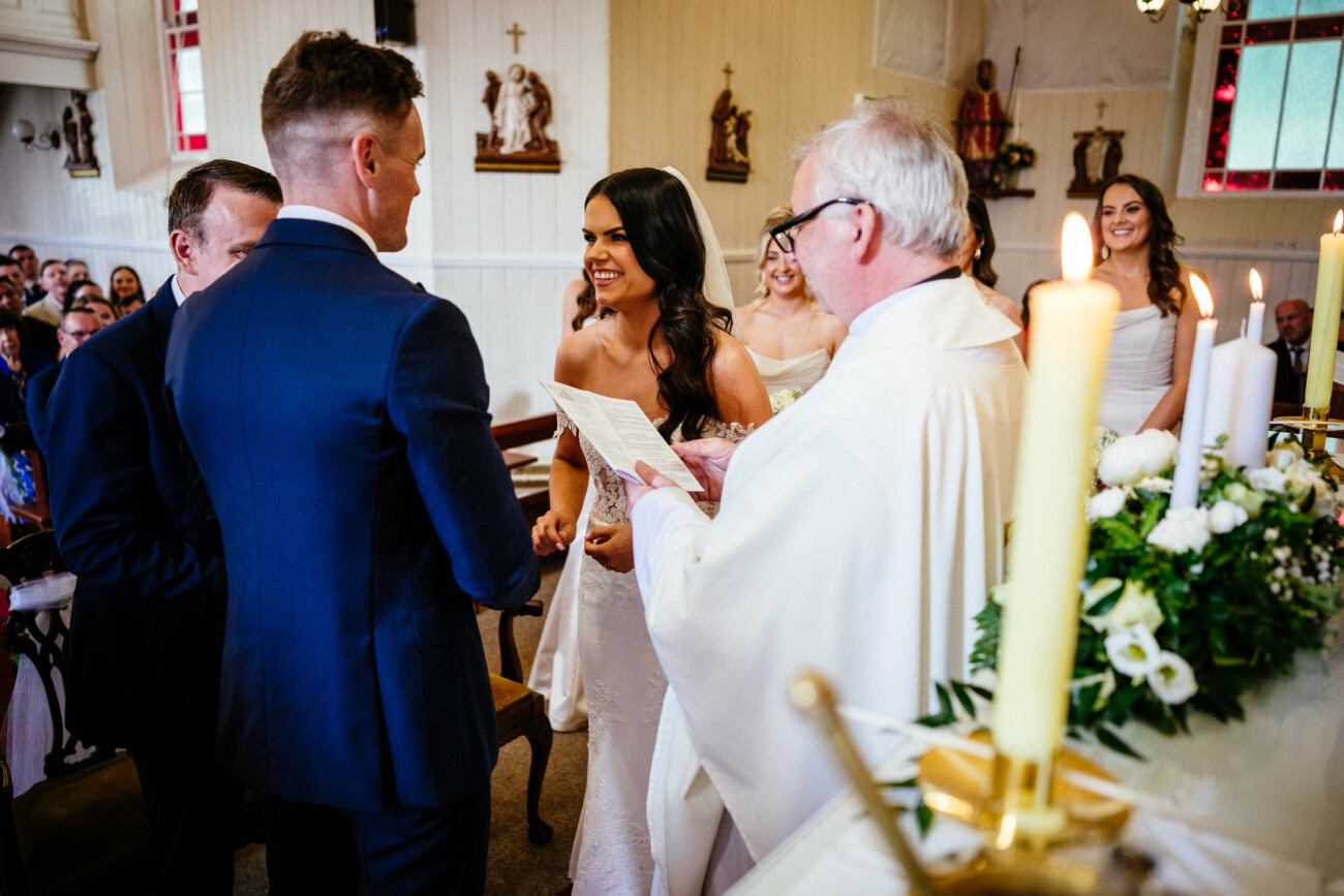 Tara and Darren’s Unforgettable Wedding at Bellingham Castle 76 A bride and groom stand facing each other at the altar in a church, with a priest officiating and guests seated in the background, capturing the timeless elegance of a wedding at Bellingham Castle.