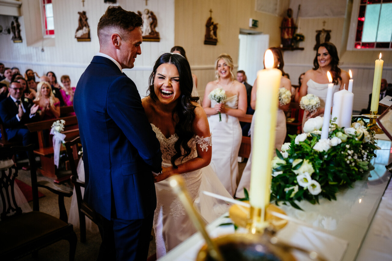 Tara and Darren’s Unforgettable Wedding at Bellingham Castle 77 A bride laughs joyfully whilst holding hands with the groom during their wedding ceremony at Bellingham Castle, surrounded by bridesmaids and guests.