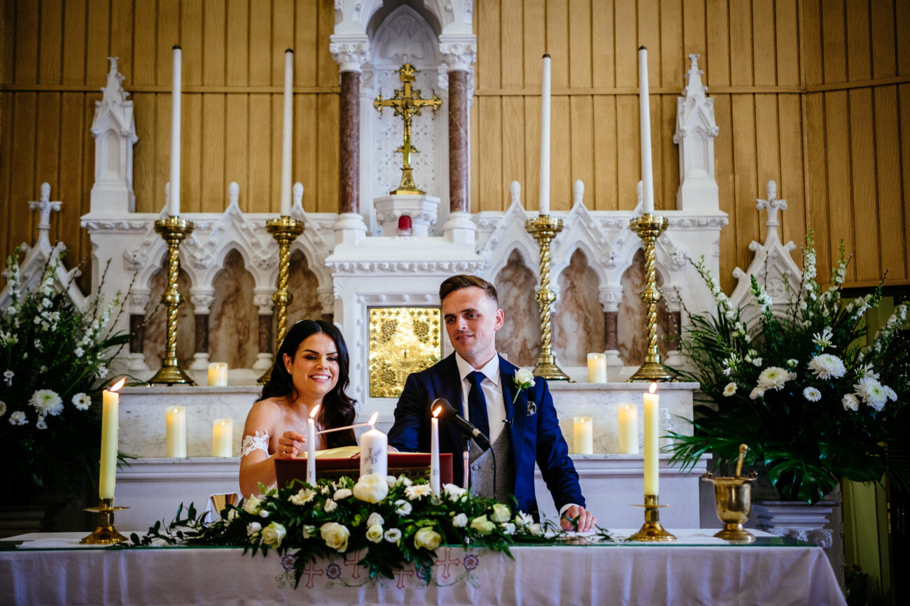 Tara and Darren’s Unforgettable Wedding at Bellingham Castle 78 A bride and groom light a candle together at an altar decorated with flowers and candles inside a church, capturing a heartfelt moment from their beautiful wedding at Bellingham Castle.