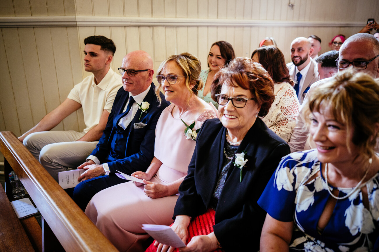 Tara and Darren’s Unforgettable Wedding at Bellingham Castle 79 Guests sit in wooden pews at a wedding at Bellingham Castle, dressed formally and smiling, holding programmes. The setting appears to be inside a church or chapel with light-coloured wooden panelling.