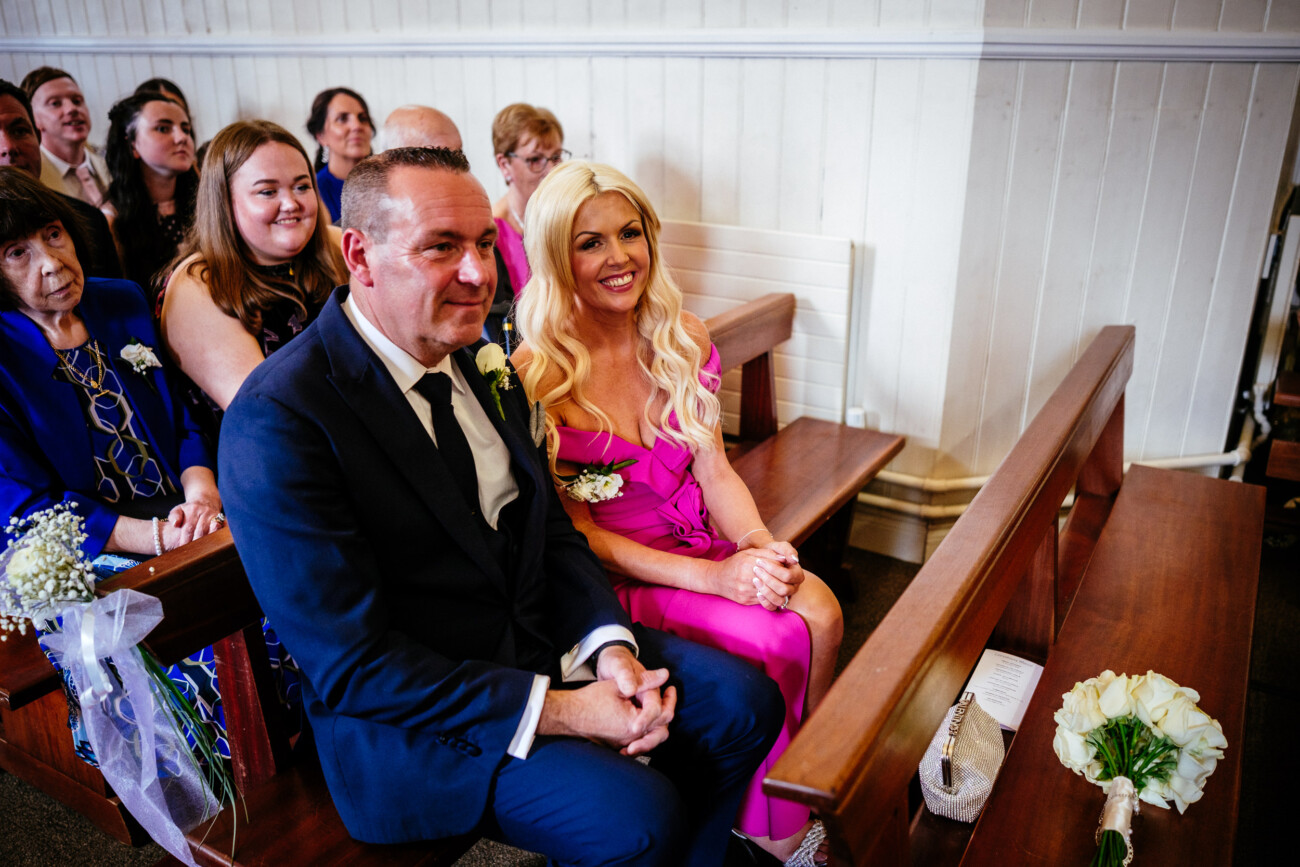 Tara and Darren’s Unforgettable Wedding at Bellingham Castle 80 A man in a suit and a woman in a pink dress sit together in a church pew, surrounded by other guests, during a beautiful wedding ceremony at Bellingham Castle.