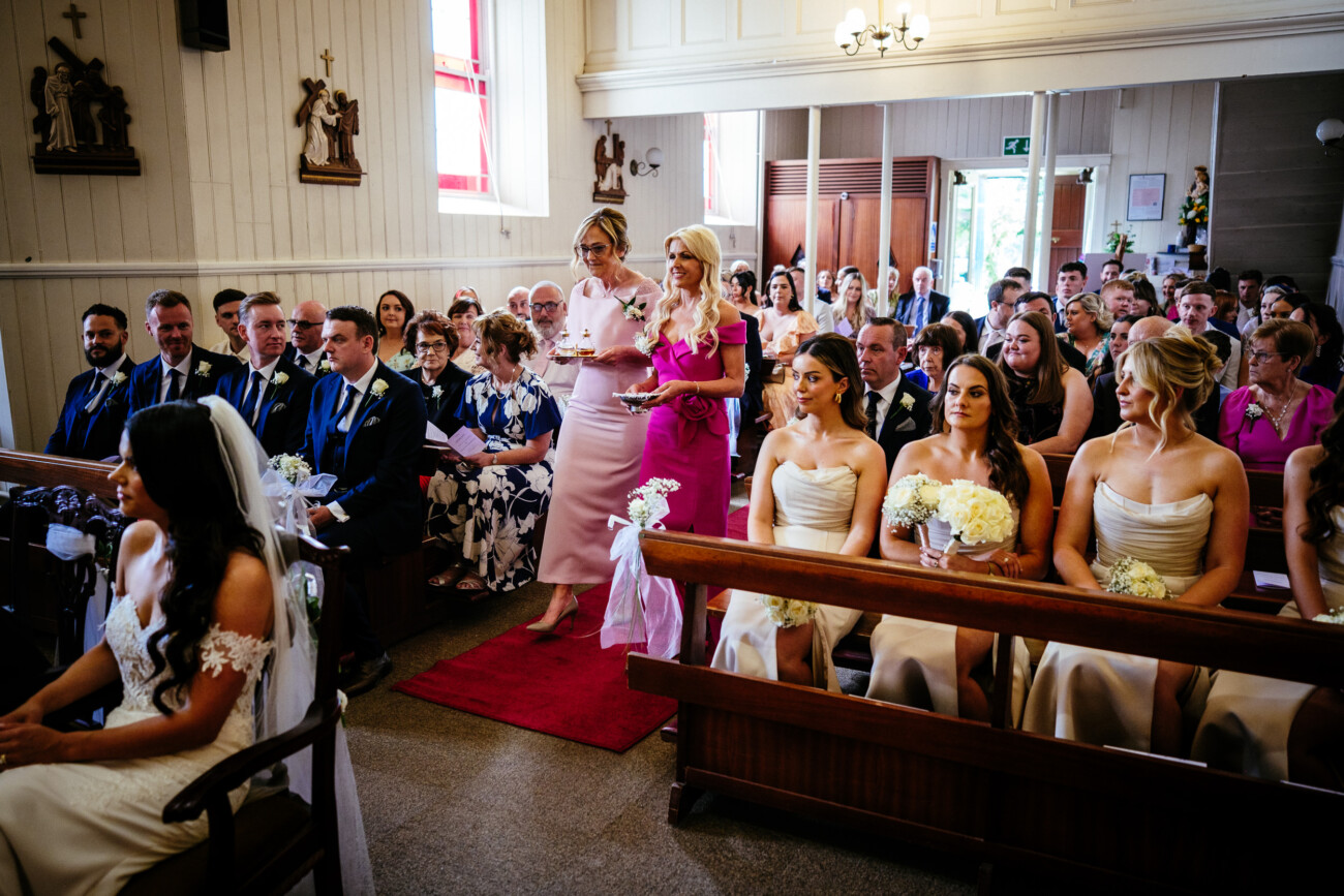 Tara and Darren’s Unforgettable Wedding at Bellingham Castle 81 Bridesmaids in pink dresses walk down the aisle of a church filled with seated guests, whilst the bride sits at the front during a beautiful wedding at Bellingham Castle.