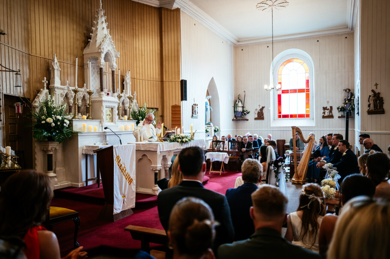 Tara and Darren’s Unforgettable Wedding at Bellingham Castle 82 A priest speaks at the altar during a wedding at Bellingham Castle, with guests seated in pews and musicians playing instruments to the side.