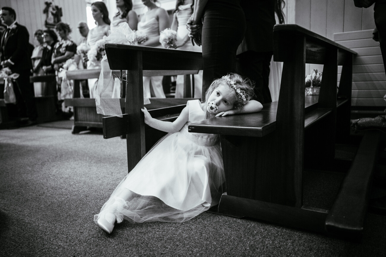 Tara and Darren’s Unforgettable Wedding at Bellingham Castle 83 A young girl in a dress and flower crown lounges on a church bench, looking tired, whilst people stand in the background during a formal event at a wedding at Bellingham Castle.
